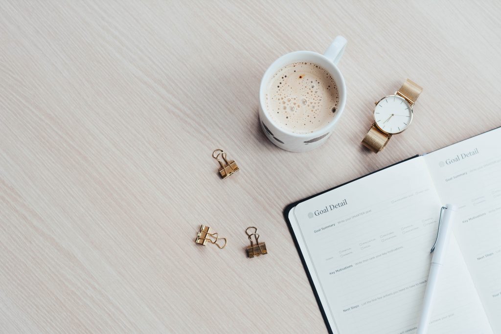 Picture of a coffee in a mug, paperclips, a watch and a blank diary wiating to be filled with healthy work habits