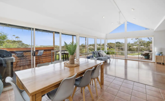 Picture of a living space in a holiday rental accommodation in Torquay that can be booked on Airbnb. The living space features floor to ceiling glass and a stunning view of greenery and ocean.