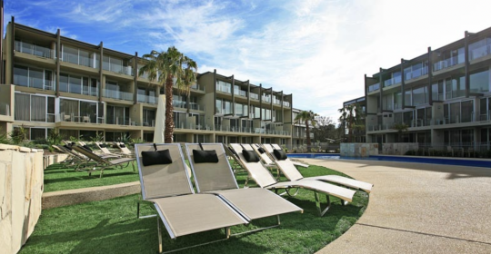 Picture of a blue sky and two buildings of apartment accommodation in Torquay, with resort beach chairs scattered on green grass in the foreground