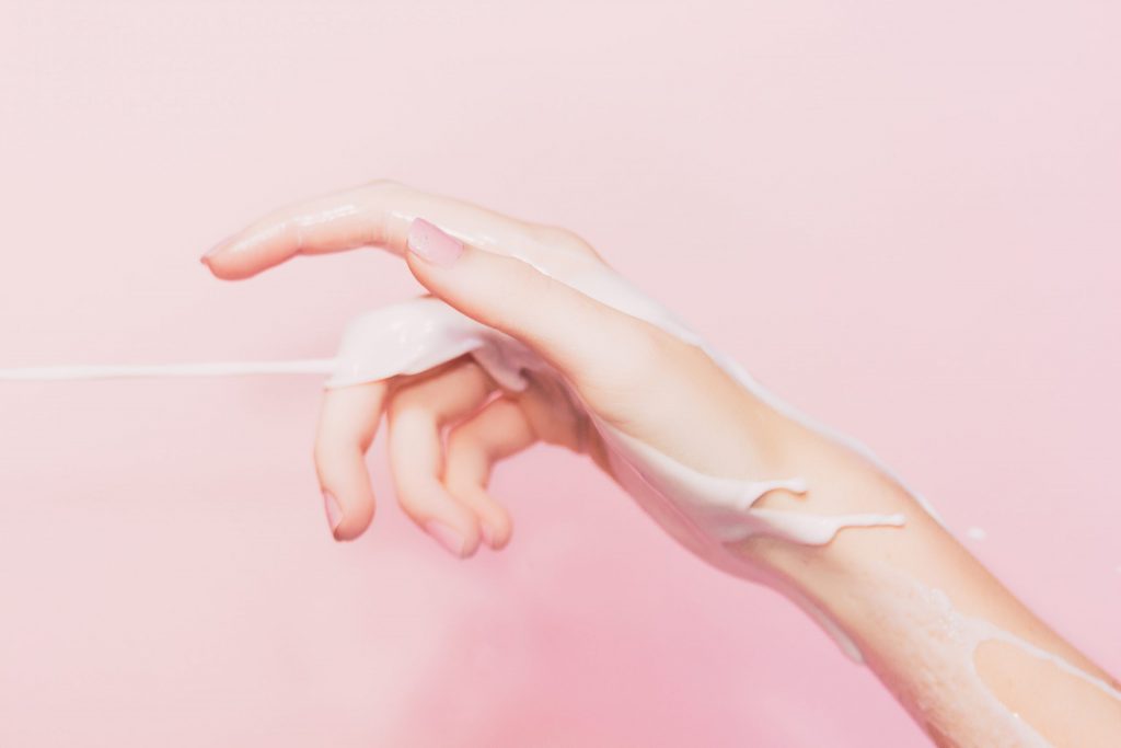 Picture of a hand with collagen balm and cleaning balms coating the skin. There is a pink background behind the hand.