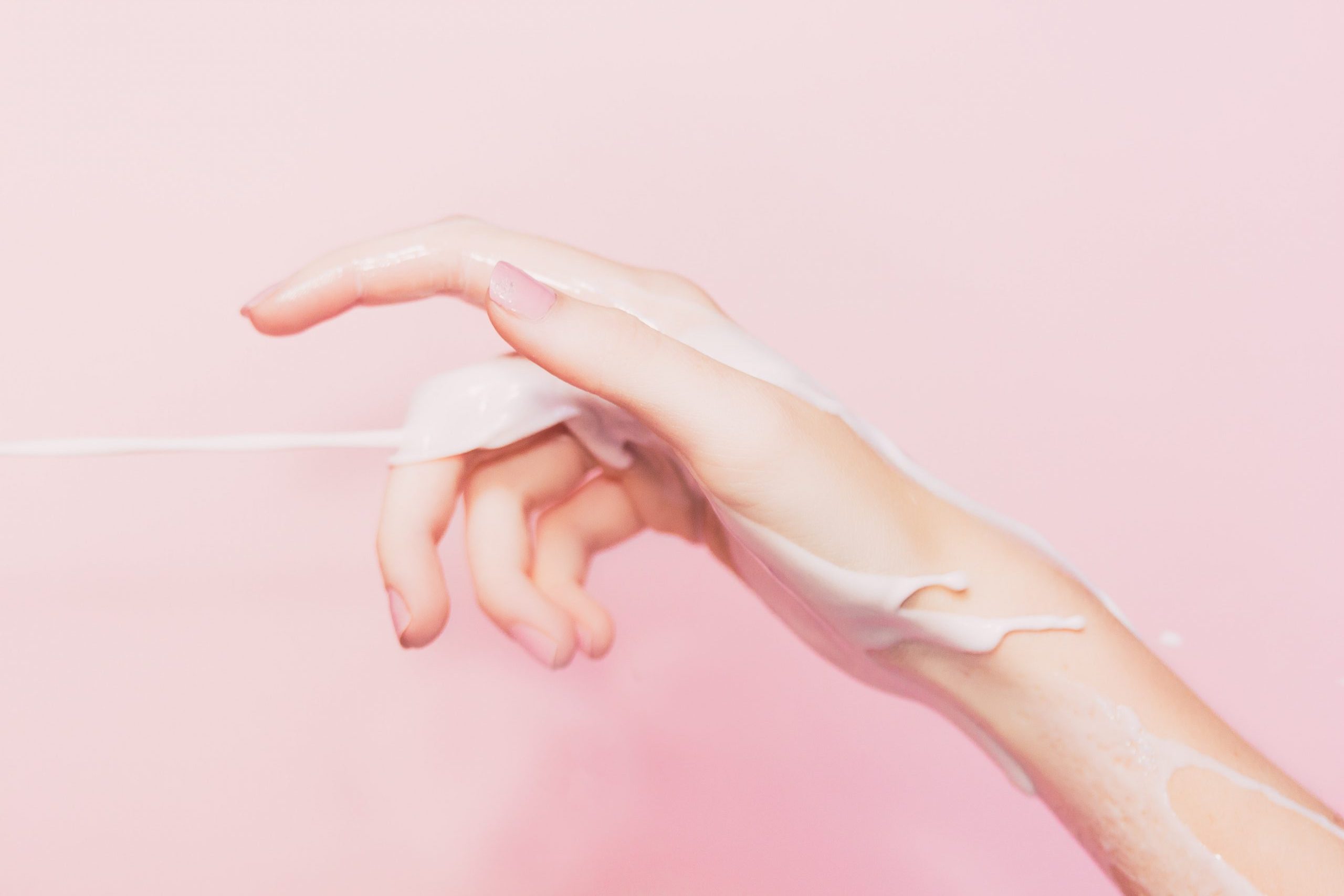 Picture of a hand with collagen balm and cleaning balms coating the skin. There is a pink background behind the hand.