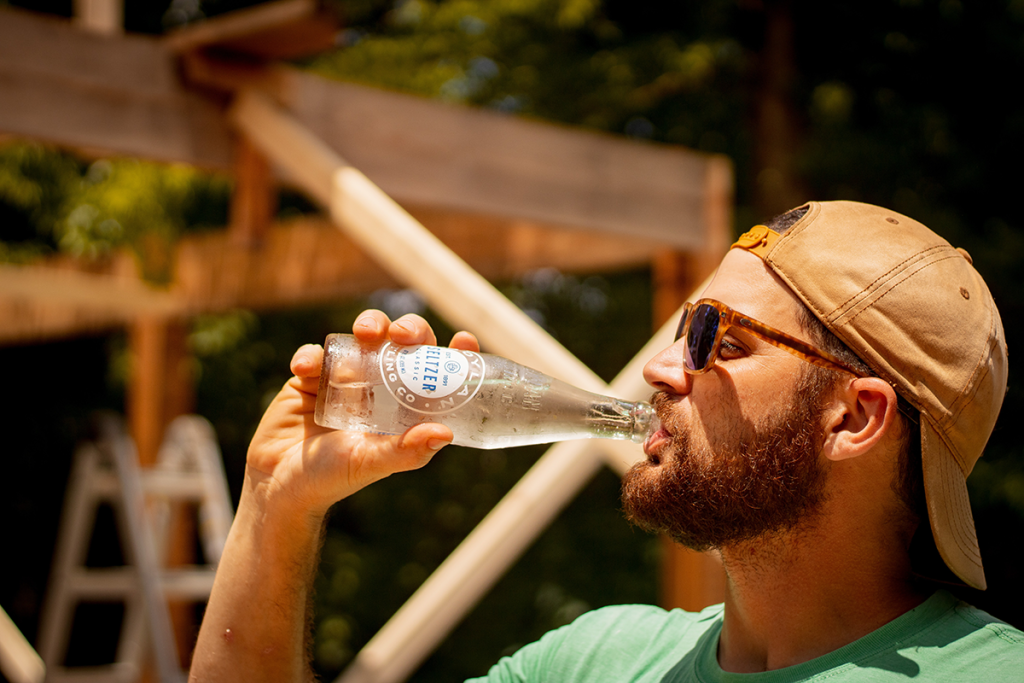 Picture of a male in a cap standing in profile drinking a glass bottle of hard seltzer on a summers day, with the seltzer bottle perspiring and glistening in the sunshine