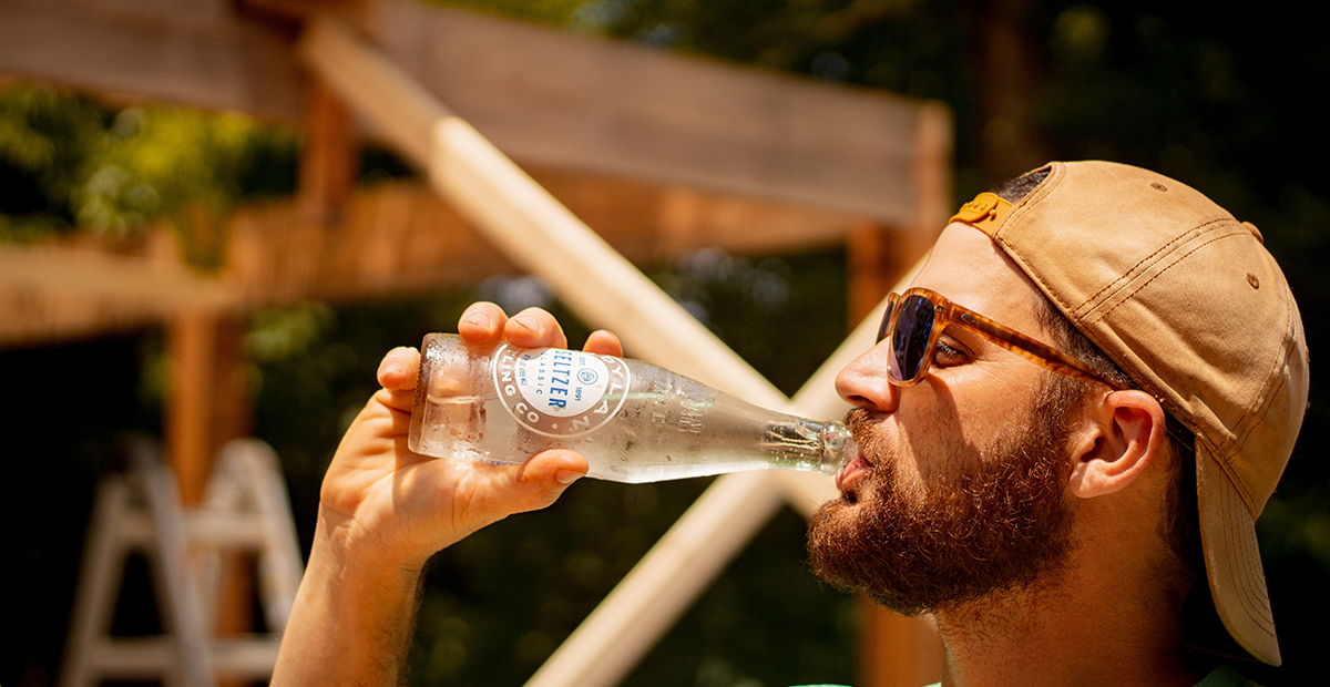 Picture of a male in a cap standing in profile drinking a glass bottle of hard seltzer on a summers day, with the seltzer bottle perspiring and glistening in the sunshine