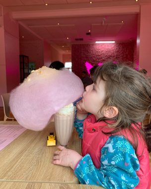 Picture of a little girl drinking a fairy floss milkshake with a big cloud of fairy floss on top of the strawberry milkshake