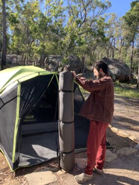 Picture of a man holding a Wanderer self inflating mattress that is rolled up, with an erected tent in the background