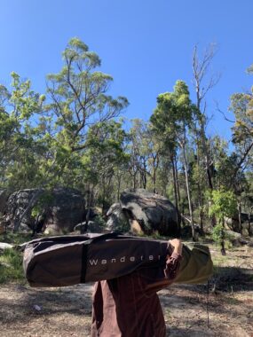 Picture of a Wanderer self inflating mattress being held over the shoulder of a man, with trees and rocks in the background and the Wanderer camping mattress logo on the bag