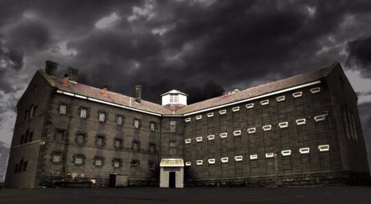 Picture of the Geelong Gaol at night time with grey clouds in the background and the upward facing photograph angle creating a towering site.