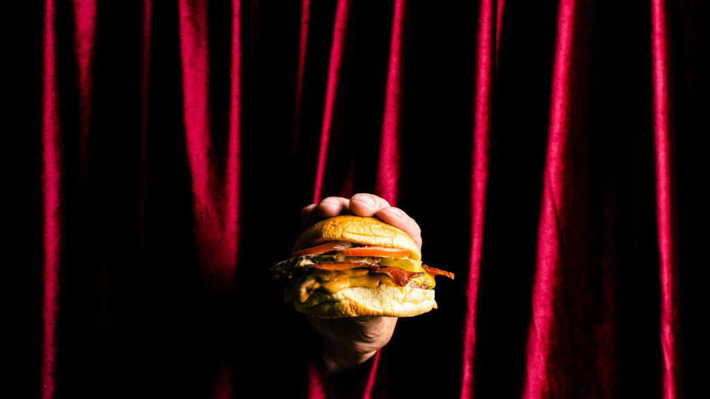 Picture of a burger being held in front of a red curtain, which is the entrance to Shush Burger