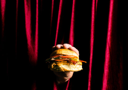 Picture of a burger being held in front of a red curtain, which is the entrance to Shush Burger