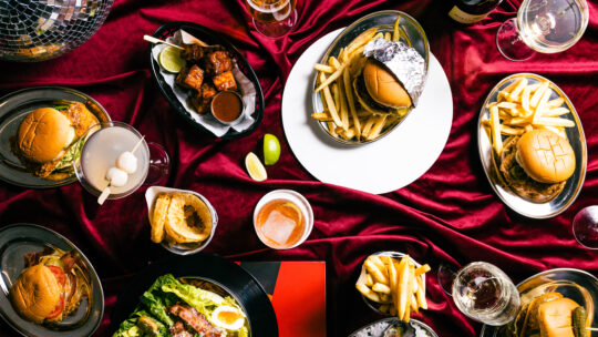 Picture of a birds-eye view of burgers, fries, cocktails, beer and dessert on a red velvet tablecloth at Shush Burger