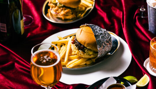 Picture of Shush Burger dishes on a velvet red tablecloth with a beer and cocktail in the foreground