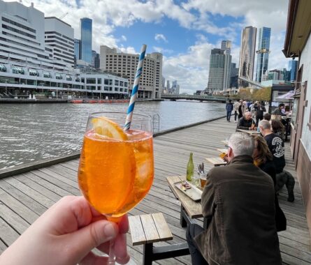 Picture of an Apreol Spritz cocktail being held against the Melbourne skyline out the front of The Boatbuilders Yard