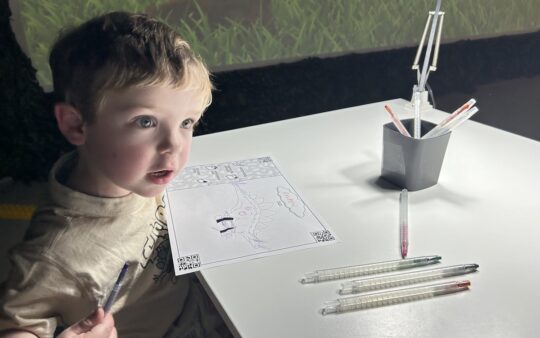 Small boy completing a colouring-in page at Dinos Alive Melbourne, which is projected onto a large screen