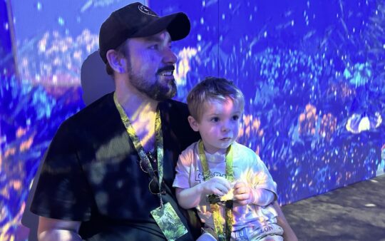 Man and small boy sitting in an underwater installation, watching underwater dinosaurs moving across the screens and floor screens