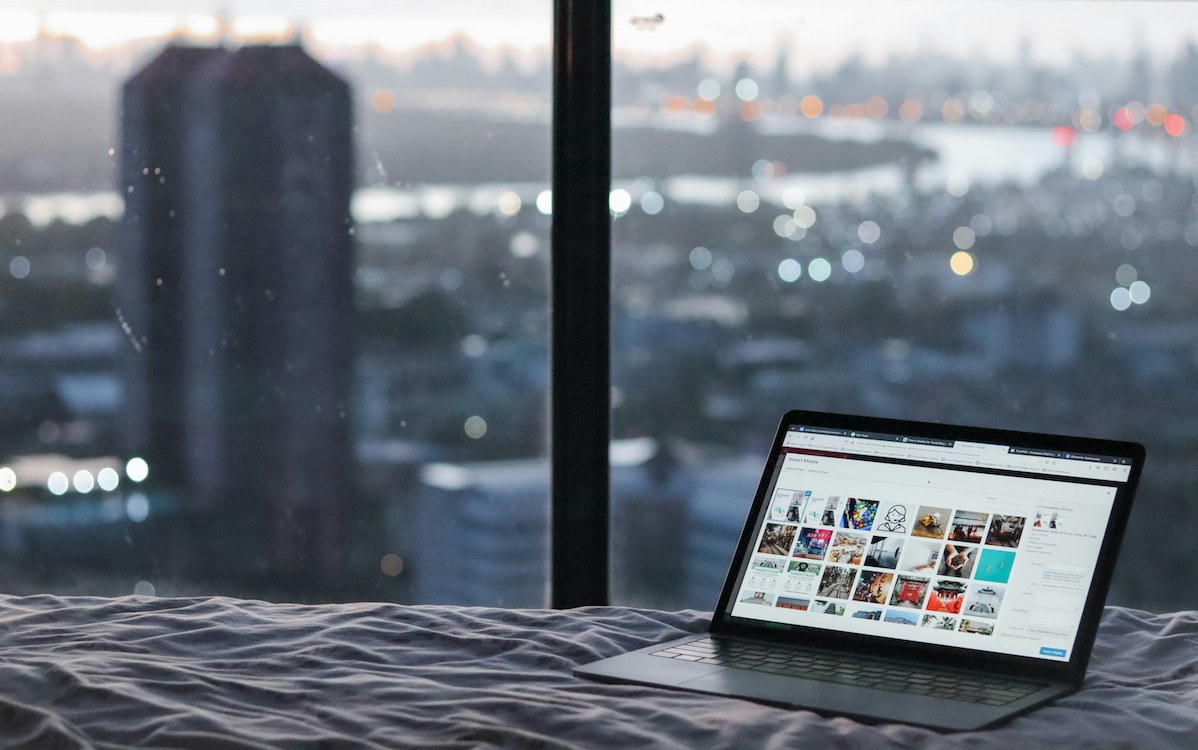 Picture of a laptop on a bed overlooking a city skyline, belonging to a digital nomad