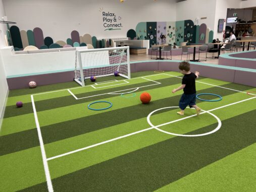 Child kicking soccer ball at an indoor soccer pitch at Eden Play Mill Park with hoops and play equipment around